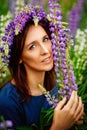 Large vertical portrait. A young brunette woman in a flower wreath holds a long lilac lupine flower near her face