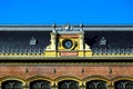 large white clock in decorative exterior yellow brick wall. black roman numerals. gray mansard zink roof