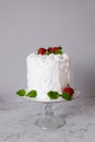 Large white delicious cake with cream and decorated with strawberries and mint on a transparent stand on a gray background