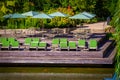 Large wooden deck by water with tables and umbrellas and green cushioned rustic rocking chairs - Lush foliage in background and