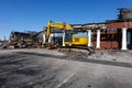 A large yellow backhoe in the parking lot in front of a building being demolished after being destroyed in a fire