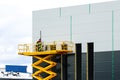 A large yellow self propelled scissor lift platform with workers against a grey sandwich panel wall