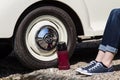 Lask sits by the wheel of a Morris Minor classic car. Legs and feet of owner wearing tennis shoes and jeans by flask