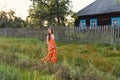 At the late in the evening a young woman in a retro village attire strolls along the old stockade and rural house