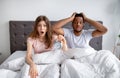 We are late. Shocked interracial couple with alarm clock grabbing their heads in terror, sitting on bed at home