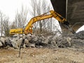 Lateral view of an excavator running a reinforced concrete demolition with a hydraulic breaker under a bridge under construction