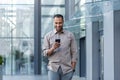 Latin american businessman in casual shirt walking near office building from outside, man smiling and using phone Royalty Free Stock Photo