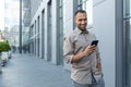 Latin american businessman in casual shirt walking near office building from outside, man smiling and using phone Royalty Free Stock Photo