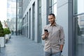 Latin american businessman in casual shirt walking near office building from outside, man smiling and using phone Royalty Free Stock Photo