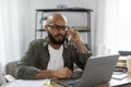 Latin male freelancer calling by cellphone while working online on laptop, having phone conversation at home Royalty Free Stock Photo