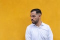 Latin man standing in front of a yellow wall. He is looking to the side with seriousness