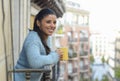 Latin woman drinking cup of coffee or tea smiling happy at apartment window balcony