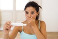 Latin woman holding pregnancy test on bed at home looking at positive result in shock and stress