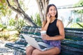 Latin pretty woman sitting on bench in green park on summer day and talking on smartphone while using silver laptop and drinking t Royalty Free Stock Photo