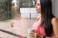 Young woman drinking a cup of coffee or tea looking out onto the terrace through a window with her image reflected in the glass