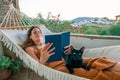 Young Hispanic woman reading a book in hammock with her cat