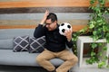 Latino adult man with glasses watches soccer game on TV on his sofa with his ball and gets angry and sad watching the football gam