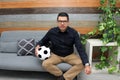 Latino adult man with glasses watches soccer game on TV on his sofa with his ball and gets excited, angry, sad watching the soccer