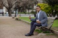 Latino man in a suit and a disposable cup of coffee sitting on a bench in a public park