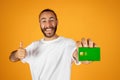 Laughing adult european man with beard in white t-shirt hold credit card and thumb up