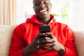 Laughing african american man sitting on couch at home using on smartphone Royalty Free Stock Photo