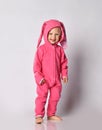 Laughing barefooted blonde baby girl in pink warm jumpsuit stands looking at camera with her hood with bunny ears on