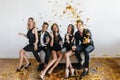 Laughing black-haired guy in black shoes throwing out golden confetti during photoshoot with friends. Indoor photo of