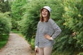 Laughing dark haired young woman standing near green leaves bush holding hand on hip wearing strip[ed shirt and panama looks happy