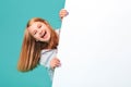 A laughing girl standing against a turquoise background holds an empty white Board in her hand and looks out from behind it Royalty Free Stock Photo