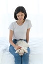Laughing jocund young asian woman sitting on bed in bedroom with her maltese dog on knees