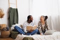 Laughing young multiethnic couple looking at each other while using laptop and reading book on bed at home Royalty Free Stock Photo