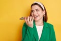 Laughing positive woman wearing green jacket posing isolated over yellow background recording voice message smiling happily copy