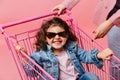 Laughing preteen kid sitting in shopping cart. Studio shot of curly child in denim jacket and sunglasses