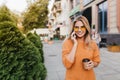 Laughing pretty lady talking on phone while walking by bushes with cup of tea. Outdoor portrait of ecstatic blonde woman