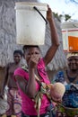 Laughing proud young African woman collecting water in buckets with sleeping albino child in a capulana on hip