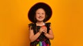 Smiling girl in Halloween witch costume holding burning candle or ignited candle and looking up on yellow background. Royalty Free Stock Photo