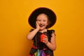 Laughing smiling girl in witch costume holding hand on cheek and holding red burning candle on yellow background. Royalty Free Stock Photo