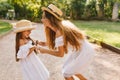 Laughing tanned woman bent over to smiling girl and dancing with her. Outdoor portrait of cute young mother and blissful