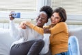 Laughing woman friends hugging each other on sofa while taking selfie photo on smart phone. Lovable caucasian and African American Royalty Free Stock Photo