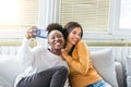 Laughing woman friends hugging each other on sofa while taking selfie photo on smart phone. Lovable caucasian and African American Royalty Free Stock Photo