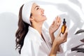 Laughing woman in a white bathrobe takes care of her skin and moisturizes it her face with a serum. Positive young female enjoys a