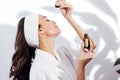 Laughing woman in a white bathrobe takes care of her skin and moisturizes it her face with a serum