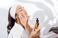 Laughing woman in a white bathrobe takes care of her skin and moisturizes it her face with a serum