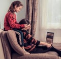 Laughing woman working at home on computer and cute kid play with mother. Sitting on sofa, daughter covering mother the eyes with Royalty Free Stock Photo