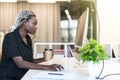 Laughing young African female entrepreneur drinking a cup of coffee and working on a laptop while sitting at a desk in office Royalty Free Stock Photo