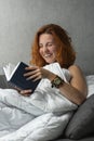 Laughing young ginger girl on the sofa under blanket reads book. Happy resting woman