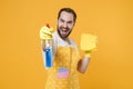 Laughing young man househusband in apron rubber gloves hold spray with washing cleanser, cleaning rag while doing