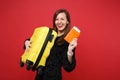 Laughing young woman in black fur sweater holding suitcase, passport and boarding pass ticket isolated on red wall Royalty Free Stock Photo