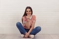 Laughing young woman covering mouth, sitting cross-legged on floor, against white brick wall. Striped shirt, jeans. Positive human Royalty Free Stock Photo