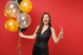 Laughing young woman in little black dress celebrating, holding glass of champagne, air balloons isolated on red Royalty Free Stock Photo